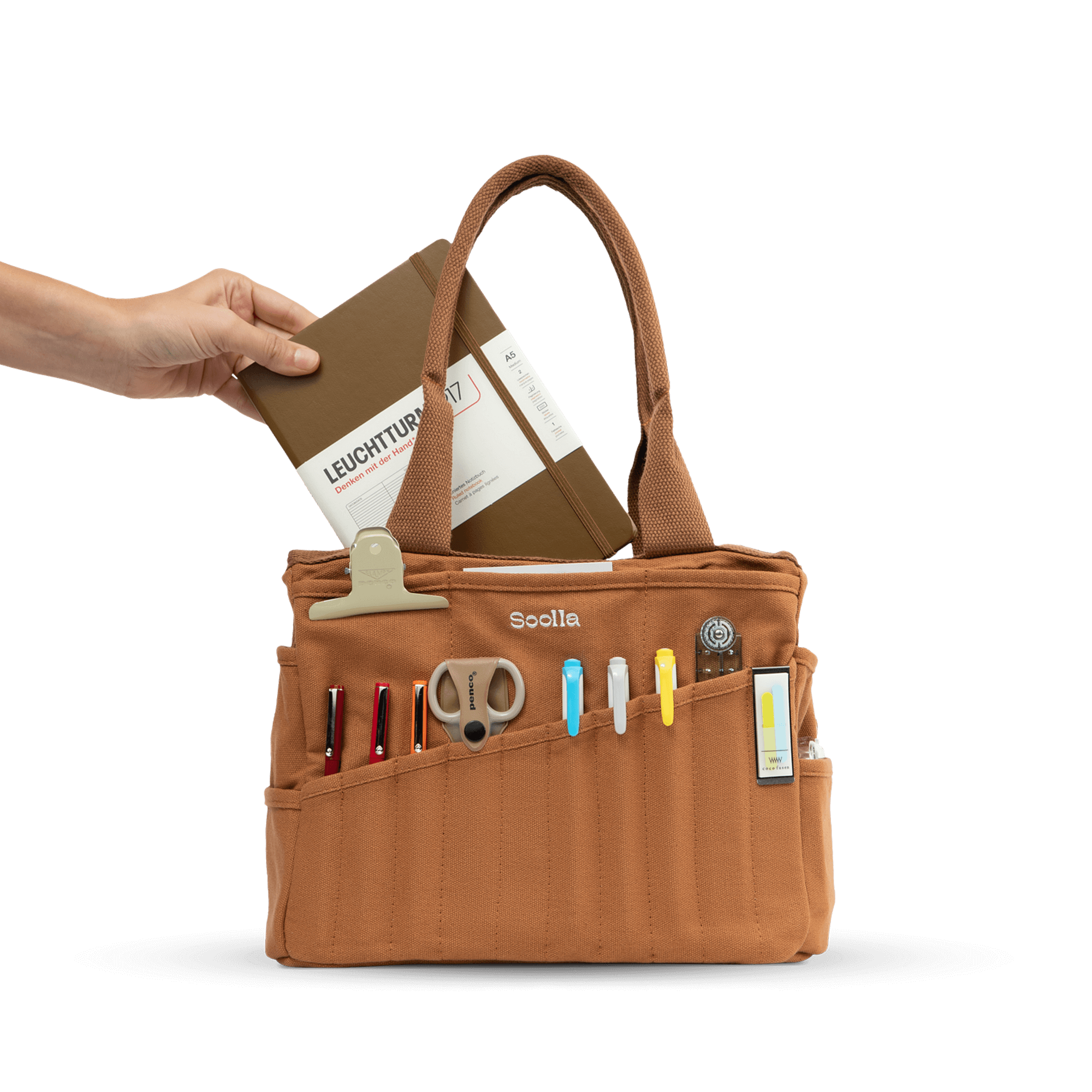 A hand is holding a brown leather tote bag with various office supplies and a notebook inside.