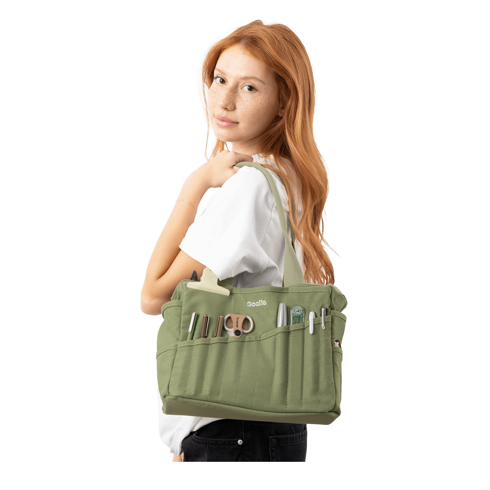 A young woman with red hair is standing against a black background, wearing a white shirt and carrying a green bag with various items in it.
